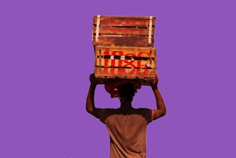 A person is carrying two stacked wooden crates on their head against a solid purple background. The crates appear worn and have red markings on them, suggesting use for transport or storage.