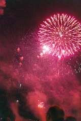 A vibrant display of pink fireworks explodes against a dark night sky. Bright bursts and trails of light create a dazzling pattern, illuminating the surrounding smoke. Silhouettes of spectators are visible in the foreground, suggesting a festive atmosphere.
