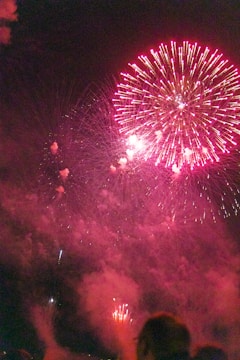 A vibrant display of pink fireworks explodes against a dark night sky. Bright bursts and trails of light create a dazzling pattern, illuminating the surrounding smoke. Silhouettes of spectators are visible in the foreground, suggesting a festive atmosphere.