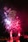 Bright, colorful fireworks explode against a dark night sky, creating vibrant patterns of red, pink, and white above a body of water. Silhouettes of people can be seen in the foreground, gazing up at the display. Puffy white clouds of smoke linger in the air, adding to the dynamic scene.