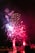 Bright, colorful fireworks explode against a dark night sky, creating vibrant patterns of red, pink, and white above a body of water. Silhouettes of people can be seen in the foreground, gazing up at the display. Puffy white clouds of smoke linger in the air, adding to the dynamic scene.