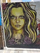 A vibrant mural on a wall depicts a woman's face with striking green eyes and long, flowing hair in shades of blonde and brown. Below her face, a stylized skull with a violin's F-holes sits surrounded by large, detailed roses in shades of pink and red.