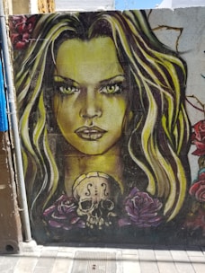 A vibrant mural on a wall depicts a woman's face with striking green eyes and long, flowing hair in shades of blonde and brown. Below her face, a stylized skull with a violin's F-holes sits surrounded by large, detailed roses in shades of pink and red.