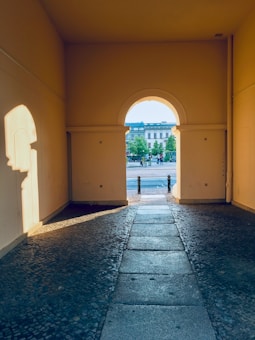 An archway leads to an outdoor area with trees and a distant building. The sunlight casts long shadows inside the archway, highlighting the textured cobblestone path. The arch frames the scene outside, creating a contrast between the dim, shadowy interior and the bright, sunlit exterior.