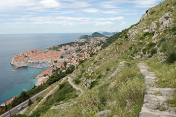 A scenic coastal view featuring a historic city with orange-roofed buildings nestled along the waterfront. The city is surrounded by ancient walls and overlooks the deep blue sea. In the foreground, a lush green hillside with rocks and a pathway descends towards the town. The sky is partly cloudy, adding to the picturesque landscape.