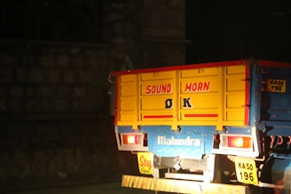 The rear portion of a brightly colored truck is parked or moving at night. The truck has a message painted on the back panel saying 'SOUND HORN OK' in bold letters. It features red, yellow, and blue colors prominently. The truck's make, Mahindra, is visible, with license plates on display. The background is dark, suggesting nighttime.