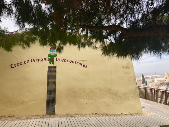 A painted wall features the phrase 'Cree en la magia y la encontrar&aacute;s' in Spanish, with a small whimsical character in a green outfit. A tree branch with green leaves hangs across the top, providing shade. The wall is a muted yellow color with a narrow vertical window or grate. A wooden platform or walkway is visible at the bottom, and a distant cityscape can be seen in the background.
