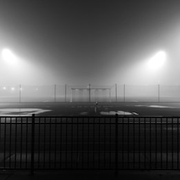 A foggy sports field with a single goalpost in the middle, illuminated by bright floodlights. There is a fence in the foreground, and the field appears empty and quiet.