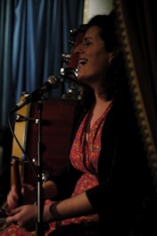 A person with curly hair, wearing a colorful floral dress, is seated and singing into a microphone. There is a drum set in the background, and a curtain visible behind them, suggesting a performance or intimate event setting. The lighting is dim, creating a cozy ambiance.