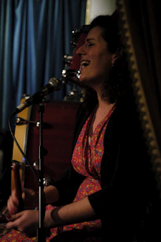 A person with curly hair, wearing a colorful floral dress, is seated and singing into a microphone. There is a drum set in the background, and a curtain visible behind them, suggesting a performance or intimate event setting. The lighting is dim, creating a cozy ambiance.