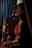 A person with curly hair, wearing a colorful floral dress, is seated and singing into a microphone. There is a drum set in the background, and a curtain visible behind them, suggesting a performance or intimate event setting. The lighting is dim, creating a cozy ambiance.