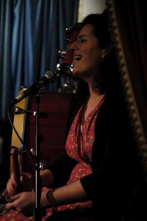 A person with curly hair, wearing a colorful floral dress, is seated and singing into a microphone. There is a drum set in the background, and a curtain visible behind them, suggesting a performance or intimate event setting. The lighting is dim, creating a cozy ambiance.