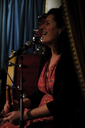 A person with curly hair, wearing a colorful floral dress, is seated and singing into a microphone. There is a drum set in the background, and a curtain visible behind them, suggesting a performance or intimate event setting. The lighting is dim, creating a cozy ambiance.