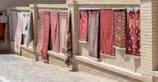 Several decorative rugs are displayed hanging outdoors against a brick wall. The rugs feature intricate designs and patterns, primarily in red and brown tones. The setting appears to be a sunlit marketplace or alley with a cobblestone path next to the wall.