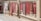 Several decorative rugs are displayed hanging outdoors against a brick wall. The rugs feature intricate designs and patterns, primarily in red and brown tones. The setting appears to be a sunlit marketplace or alley with a cobblestone path next to the wall.