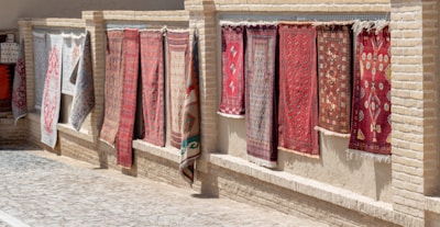 Several decorative rugs are displayed hanging outdoors against a brick wall. The rugs feature intricate designs and patterns, primarily in red and brown tones. The setting appears to be a sunlit marketplace or alley with a cobblestone path next to the wall.