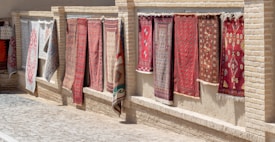 Several decorative rugs are displayed hanging outdoors against a brick wall. The rugs feature intricate designs and patterns, primarily in red and brown tones. The setting appears to be a sunlit marketplace or alley with a cobblestone path next to the wall.