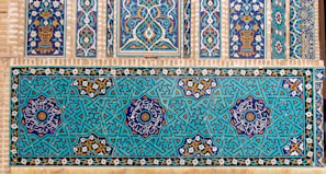 A detailed display of intricate, geometric Islamic tiles with floral motifs. The tiles feature complex interlocking patterns in a vibrant teal and navy color scheme, accented with white and gold detailing. Horizontal and vertical panels showcase symmetrical designs incorporating stars and floral symbols.