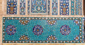 A detailed display of intricate, geometric Islamic tiles with floral motifs. The tiles feature complex interlocking patterns in a vibrant teal and navy color scheme, accented with white and gold detailing. Horizontal and vertical panels showcase symmetrical designs incorporating stars and floral symbols.