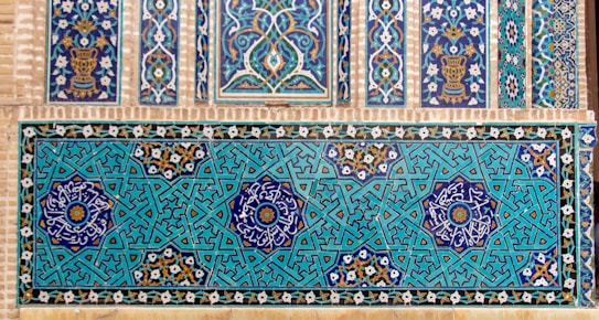 A detailed display of intricate, geometric Islamic tiles with floral motifs. The tiles feature complex interlocking patterns in a vibrant teal and navy color scheme, accented with white and gold detailing. Horizontal and vertical panels showcase symmetrical designs incorporating stars and floral symbols.