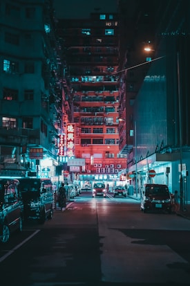 A nighttime urban scene featuring a narrow street lined with tall, densely packed apartment buildings. Bright neon signs in red and blue illuminate the area, casting colorful reflections on the parked cars and pavement. The street is relatively quiet with a few vehicles and a couple of pedestrians visible.