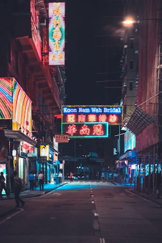 A dimly lit urban street with vibrant neon signs in various colors creating an atmospheric nighttime scene. Pedestrians stroll along the sidewalks beside storefronts, with the largest sign reading 'Koon Nam Wah Bridal'. The environment is bustling yet calm, capturing the essence of a city at night.