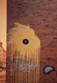 A brick wall features a large mural with dripping yellow paint that forms an abstract design. At the center of the mural is a circular black shape with an orange dot in the middle, resembling an eye. Below the artwork, there are layers of graffiti in various forms and colors, including black and blue scribbles.
