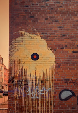 A brick wall features a large mural with dripping yellow paint that forms an abstract design. At the center of the mural is a circular black shape with an orange dot in the middle, resembling an eye. Below the artwork, there are layers of graffiti in various forms and colors, including black and blue scribbles.