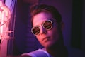 A person wearing round sunglasses reflects neon lights in their lenses. The ambient lighting creates a moody atmosphere with predominant purple and pink hues, giving a futuristic and urban vibe.