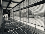 A modern corridor with large windows is characterized by horizontal blinds casting intricate shadows on the floor. The space is empty, and the blinds allow partial views of the outside urban environment.