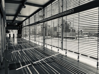 A modern corridor with large windows is characterized by horizontal blinds casting intricate shadows on the floor. The space is empty, and the blinds allow partial views of the outside urban environment.