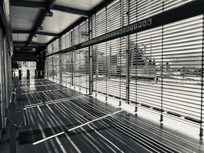 A modern corridor with large windows is characterized by horizontal blinds casting intricate shadows on the floor. The space is empty, and the blinds allow partial views of the outside urban environment.