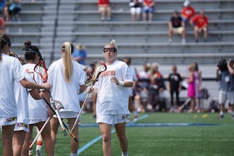 A group of lacrosse players in white uniforms gather on a green field while holding lacrosse sticks. They appear to be preparing for a game or practice. Spectators and a cameraman are visible in the background, seated on metal bleachers.