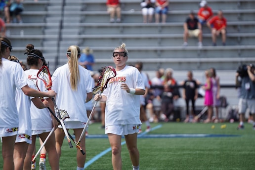 A group of lacrosse players in white uniforms gather on a green field while holding lacrosse sticks. They appear to be preparing for a game or practice. Spectators and a cameraman are visible in the background, seated on metal bleachers.