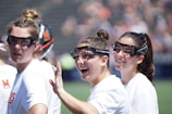 Three women with goggles stand closely together, wearing white sports shirts with logos on them. One woman waves toward the camera with a wide smile, adding a sense of camaraderie among them. The background is out of focus, possibly a sports field, indicating an outdoor setting.