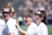 Three women with goggles stand closely together, wearing white sports shirts with logos on them. One woman waves toward the camera with a wide smile, adding a sense of camaraderie among them. The background is out of focus, possibly a sports field, indicating an outdoor setting.