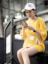 A young woman is sitting outdoors wearing a casual outfit with a yellow oversized t-shirt and matching bag. She is focused on her smartphone, wearing a white cap with a cat graphic and large hoop earrings.