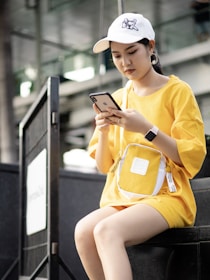 A young woman is sitting outdoors wearing a casual outfit with a yellow oversized t-shirt and matching bag. She is focused on her smartphone, wearing a white cap with a cat graphic and large hoop earrings.