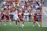 Athletes engaged in a competitive lacrosse match on a grassy field, with one player in white attempting to score against a goalie in burgundy gear. The scene is lively, with a full audience in the background, dressed casually and attentively watching the game.