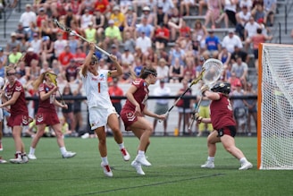 Athletes engaged in a competitive lacrosse match on a grassy field, with one player in white attempting to score against a goalie in burgundy gear. The scene is lively, with a full audience in the background, dressed casually and attentively watching the game.