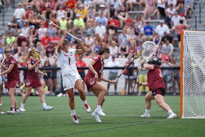 Athletes engaged in a competitive lacrosse match on a grassy field, with one player in white attempting to score against a goalie in burgundy gear. The scene is lively, with a full audience in the background, dressed casually and attentively watching the game.
