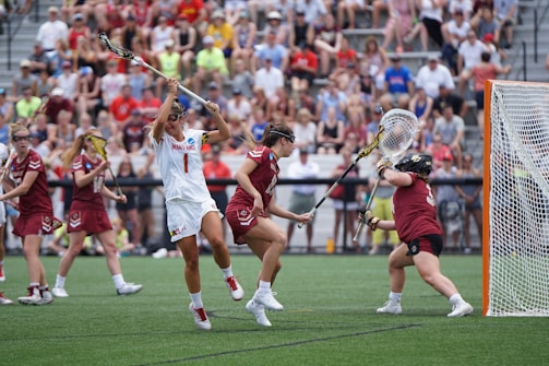 Athletes engaged in a competitive lacrosse match on a grassy field, with one player in white attempting to score against a goalie in burgundy gear. The scene is lively, with a full audience in the background, dressed casually and attentively watching the game.