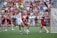 Athletes engaged in a competitive lacrosse match on a grassy field, with one player in white attempting to score against a goalie in burgundy gear. The scene is lively, with a full audience in the background, dressed casually and attentively watching the game.