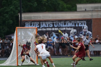 A lacrosse game in progress with a player attempting to score a goal while being closely defended by opponents. The goalkeeper stands ready to block the shot. The scene takes place on a field surrounded by a crowd of spectators, with a large banner in the background reading 'BLUE JAYS UNLIMITED.' A referee is visible nearby.