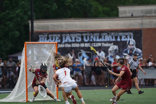 A lacrosse game in progress with a player attempting to score a goal while being closely defended by opponents. The goalkeeper stands ready to block the shot. The scene takes place on a field surrounded by a crowd of spectators, with a large banner in the background reading 'BLUE JAYS UNLIMITED.' A referee is visible nearby.