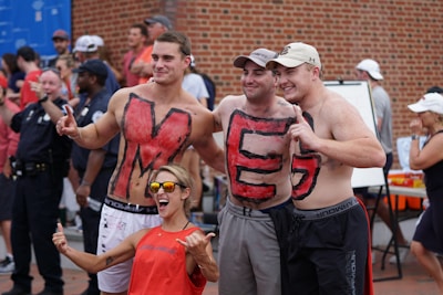 A group of three men pose together, shirtless, with painted letters 'M', 'E', and 'G' on their chests. They are standing close to each other, smiling and gesturing with peace signs. A woman in front of them, wearing sunglasses and a red tank top, is enthusiastically posing with a hand gesture. In the background, a crowd of people, including police officers, can be seen.