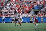 A sports event with two athletes engaged in a lacrosse game. One athlete in a white jersey is holding a lacrosse stick, appearing to prepare for a play. A referee in a striped uniform is nearby. The background features a large audience seated in a stadium, with many spectators wearing bright and casual clothing. Some photographers are also visible near the field.