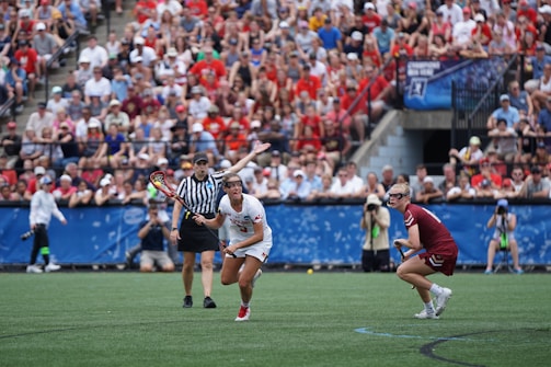 A sports event with two athletes engaged in a lacrosse game. One athlete in a white jersey is holding a lacrosse stick, appearing to prepare for a play. A referee in a striped uniform is nearby. The background features a large audience seated in a stadium, with many spectators wearing bright and casual clothing. Some photographers are also visible near the field.
