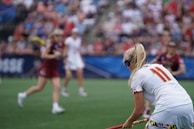 A sporting event with athletes on a grass field, one in a white jersey with the number 11 in the foreground, holding a lacrosse stick. Players in the background wear maroon uniforms. Spectators fill the stands creating a lively atmosphere.