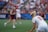 A sporting event with athletes on a grass field, one in a white jersey with the number 11 in the foreground, holding a lacrosse stick. Players in the background wear maroon uniforms. Spectators fill the stands creating a lively atmosphere.